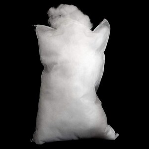 Op de afbeelding: Een witte, rechthoekige zak gevuld met zachte, witte vulling. De zak is gemaakt van een non-woven stof en is aan de bovenkant open, waardoor meer van de zachte vulling zichtbaar is. De achtergrond is zwart.