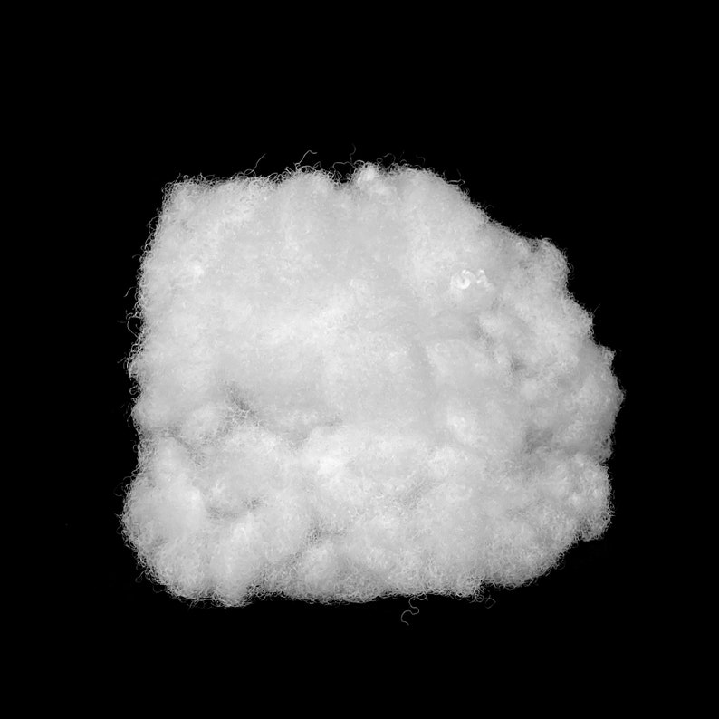 Op de afbeelding: Een close-up van een massa wit, pluizig materiaal, dat op een wolk lijkt, tegen een zwarte achtergrond. Het materiaal heeft een textuur en een onregelmatige vorm. De vezels zijn dicht opeengepakt, waardoor het een volumineuze uitstraling heeft, mogelijk gebruikt voor vulling of opvulling.