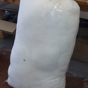 Op de afbeelding: Een grote, witte zak gevuld met een zacht materiaal, verzegeld met doorzichtig plastic. Handgeschreven markeringen zijn zichtbaar op de zak. De zak staat rechtop op een vloer.
