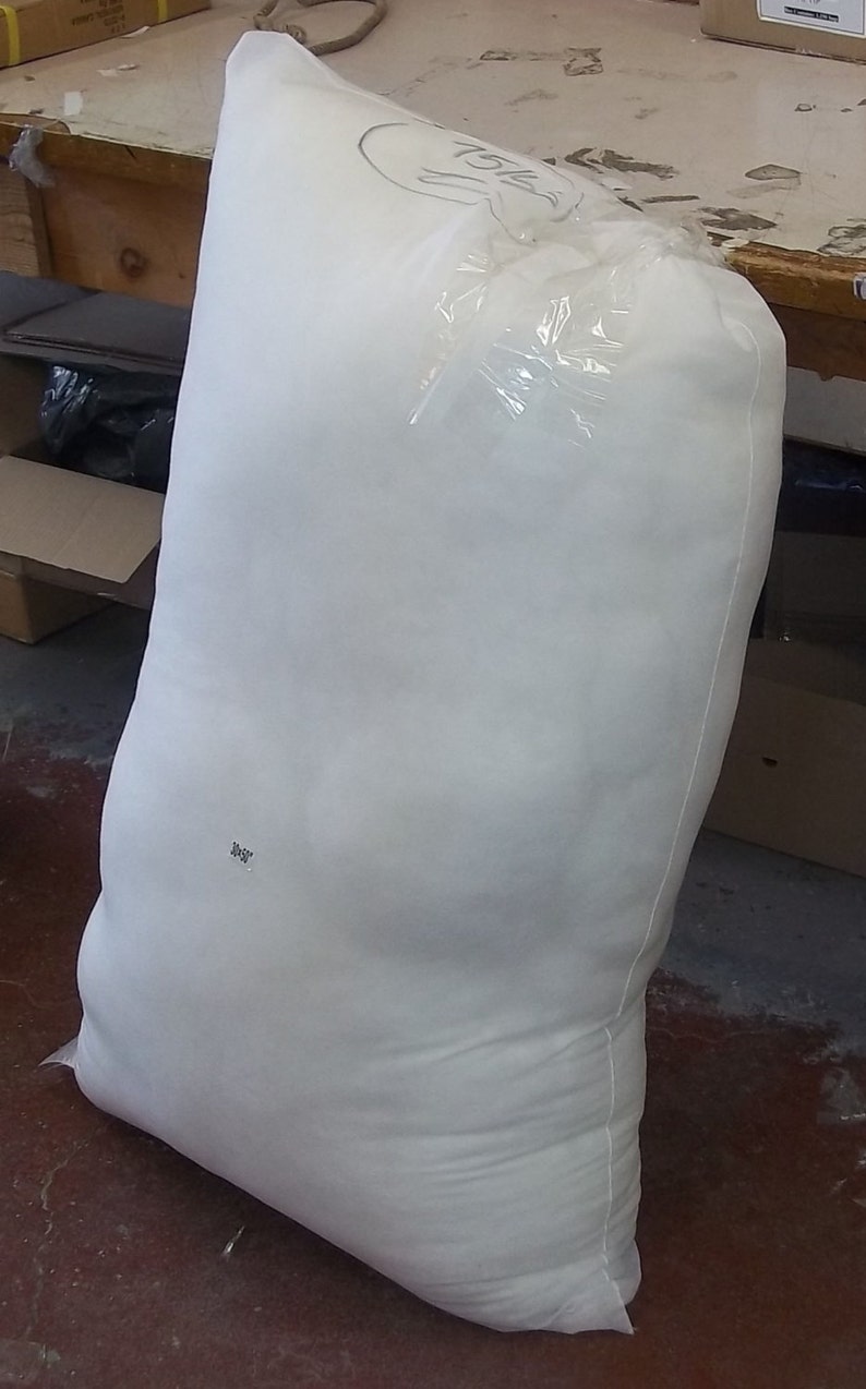 Op de afbeelding: Een grote, witte zak gevuld met een zacht materiaal, verzegeld met doorzichtig plastic. Handgeschreven markeringen zijn zichtbaar op de zak. De zak staat rechtop op een vloer.