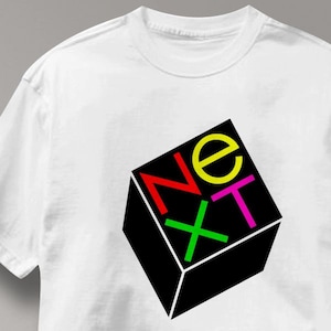 May include: White t-shirt featuring a black cube with the word "NEXT" in red, yellow, green, and pink. The design is a bold, geometric graphic, perfect for casual wear.