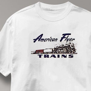 May include: White t-shirt featuring a vintage illustration of a steam train with the text "American Flyer Trains" in navy blue. The train is black and white with red train cars.