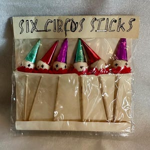 May include: A pack of six circus sticks. Each stick has a small clown head with a colourful cone hat in green, red, and purple. The heads sit on a red base with white trim. The package has the text "SIX CIRCUS STICKS".