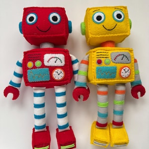Op de afbeelding: Twee handgemaakte vilten robotspeelgoed. Eén is rood met blauw-wit gestreepte armen en benen, de andere is geel met grijs-oranje gestreepte armen en benen. Beide hebben lachende gezichten en meters op hun borst.
