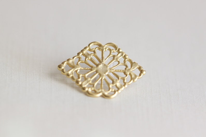 May include: A gold-colored metal decorative embellishment with a square shape and intricate, lacy design.