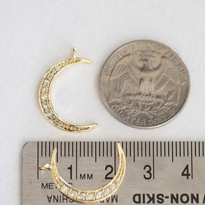 May include: Gold crescent moon charm with clear rhinestones around the edge. The charm is approximately 2.5 cm in diameter and is shown next to a US quarter for size comparison.