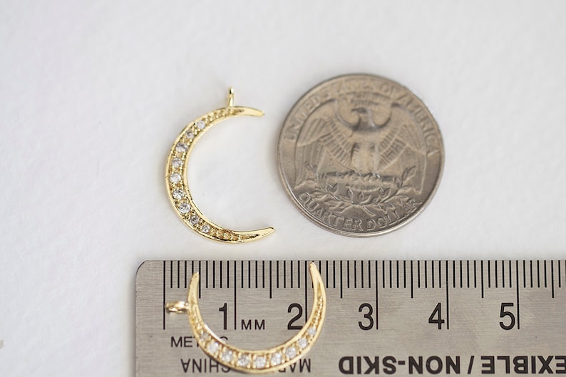 May include: Gold crescent moon charm with clear rhinestones around the edge. The charm is approximately 2.5 cm in diameter and is shown next to a US quarter for size comparison.