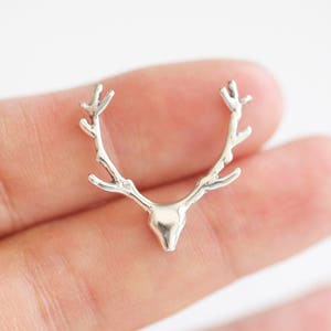 May include: A silver-colored antler charm with a detailed design. The charm is shaped like a deer's antlers and head. The charm is held in a person's hand against a white background. This charm is suitable for jewelry making.