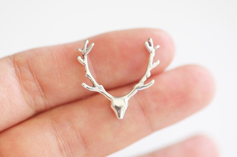 May include: A silver-colored antler charm with a detailed design. The charm is shaped like a deer's antlers and head. The charm is held in a person's hand against a white background. This charm is suitable for jewelry making.