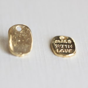 May include: Two small, gold-colored metal tags. One tag is blank, with a hole at the top. The other tag has the words "MADE WITH LOVE" embossed on it. Both tags have a slightly irregular, rectangular shape.