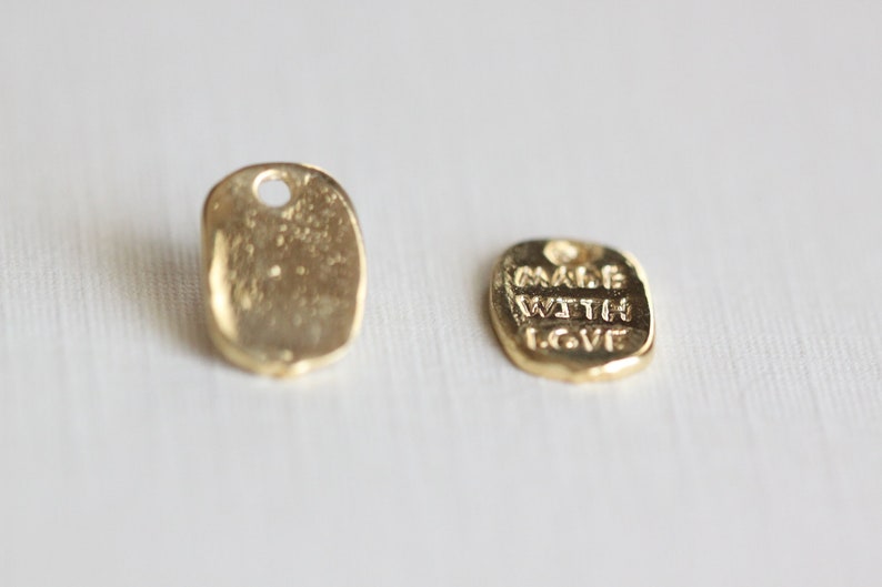 May include: Two small, gold-colored metal tags. One tag is blank, with a hole at the top. The other tag has the words "MADE WITH LOVE" embossed on it. Both tags have a slightly irregular, rectangular shape.