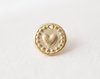 May include: A gold-colored, round charm with a heart in the center. The heart is surrounded by a beaded border. The charm has a small hole at the top for stringing.