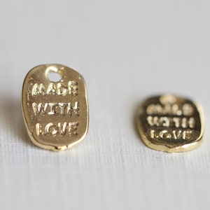 May include: Two small, gold-colored metal tags with the words "MADE WITH LOVE" embossed on them. The tags are rectangular with rounded edges and a small hole at the top for attaching to a product.