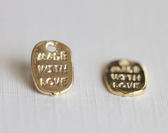 May include: Two small, gold-coloured metal tags with the words "MADE WITH LOVE" embossed on them. The tags are rectangular with rounded edges and a small hole at the top for attaching to a product.