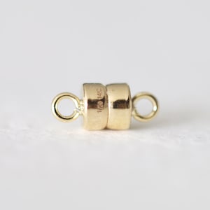 May include: A gold-colored magnetic clasp with two cylindrical sections and small loops on each end. The clasp has a shiny finish and is designed for jewelry making. The text "1/20 14K GF" is stamped on the side.