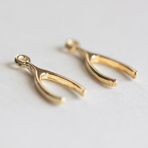 Puede incluir: Dos abalorios de color dorado en forma de hueso de la suerte. Los abalorios son pequeños y delicados, con un bucle en la parte superior para sujetarlos a un collar o pulsera.