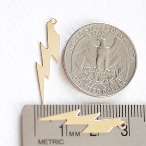 May include: Gold-colored lightning bolt charms, with a quarter for size comparison. The charms are shown with a metric ruler, and have a small hole at the top for stringing. These charms are ideal for jewelry making.