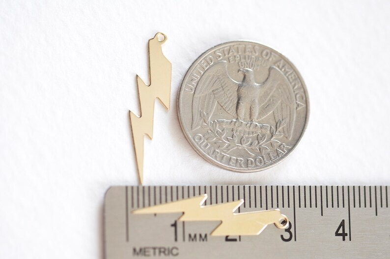 May include: Gold-colored lightning bolt charms, with a quarter for size comparison. The charms are shown with a metric ruler, and have a small hole at the top for stringing. These charms are ideal for jewelry making.