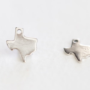 May include: Two silver charms shaped like the state of Texas. The charms have a hole at the top for attaching to a necklace or bracelet.