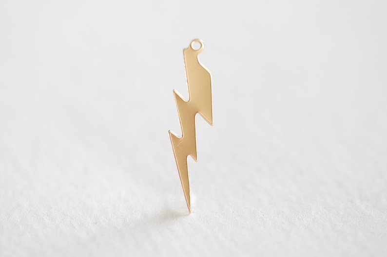 May include: A gold-colored lightning bolt charm with a small loop at the top. The charm has a jagged, zig-zag design and is likely made of metal. The lightning bolt is a popular symbol used in jewelry and accessories.