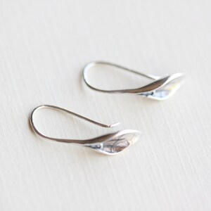 May include: A pair of silver-toned earrings. Each earring features a curved hook and a leaf-shaped design. The earrings are displayed on a light-colored surface, with a soft focus background.