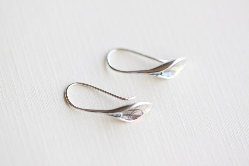May include: A pair of silver-toned earrings. Each earring features a curved hook and a leaf-shaped design. The earrings are displayed on a light-colored surface, with a soft focus background.