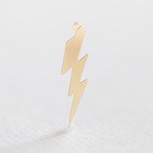May include: A gold-colored lightning bolt charm with a small loop at the top. The charm has a jagged, zig-zag design and is likely used for jewelry making or as a pendant. The background is a soft, neutral color.
