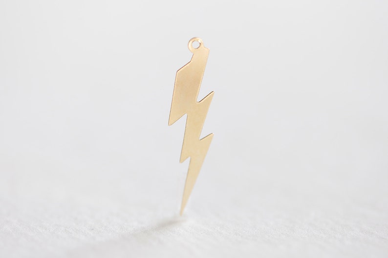 May include: A gold-colored lightning bolt charm with a small loop at the top. The charm has a jagged, zig-zag design and is likely used for jewelry making or as a pendant. The background is a soft, neutral color.