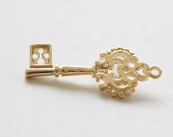 May include: A gold-toned ornate key with a decorative, intricate design. The key has a square-shaped bow and a long, slender shaft.