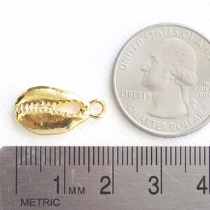 May include: Gold-colored cowrie shell charm with a loop for attaching to jewelry. The charm is about 1 centimeter long and is shown next to a US quarter for size comparison.