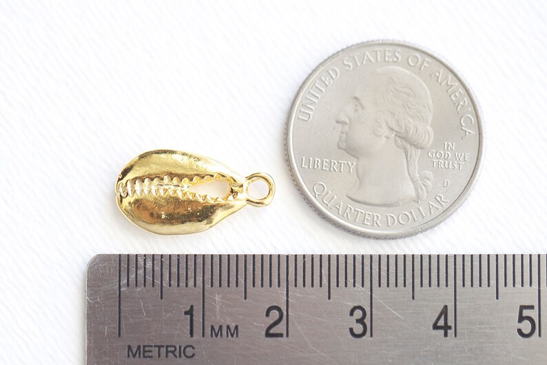 May include: Gold-colored cowrie shell charm with a loop for attaching to jewelry. The charm is about 1 centimeter long and is shown next to a US quarter for size comparison.