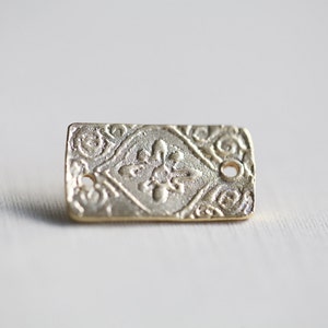 May include: A gold-colored metal charm with a decorative design and two holes. The charm is rectangular and has a raised, intricate pattern.