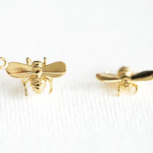 Puede incluir: Dos abalorios de abeja de oro con un bucle para sujetar a un collar o pulsera. Las abeilles están detalladas con alas, patas y un cuerpo segmentado.