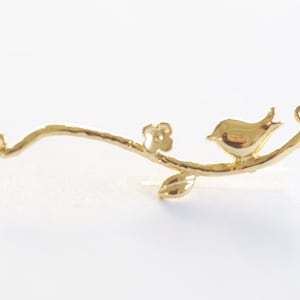 May include: Gold-toned metal charm featuring a small bird perched on a branch with a leaf and a flower. The charm has two jump rings for attaching to jewelry.