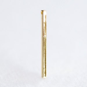 May include: A gold-colored metal bar with a textured surface. The bar has a small hole at the top.