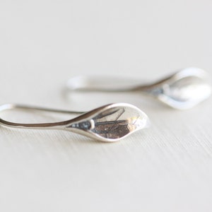 May include: A pair of silver-colored earrings. Each earring features a curved, elongated design with a petal-like shape at the end. The earrings are displayed on a light-colored surface, showcasing their reflective quality.