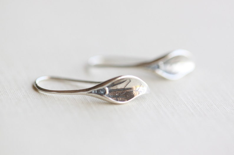 May include: A pair of silver-colored earrings. Each earring features a curved, elongated design with a petal-like shape at the end. The earrings are displayed on a light-colored surface, showcasing their reflective quality.