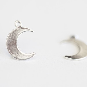 Puede incluir: Dos colgantes plateados con forma de media luna. Cada colgante tiene un pequeño lazo en la parte superior para sujetarlo a una joya. Los colgantes tienen una superficie texturizada y un aspecto ligeramente envejecido.