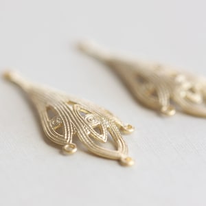 May include: Two gold-colored metal filigree embellishments with a teardrop shape and intricate designs. The embellishments have a loop at the top for attaching to a project.