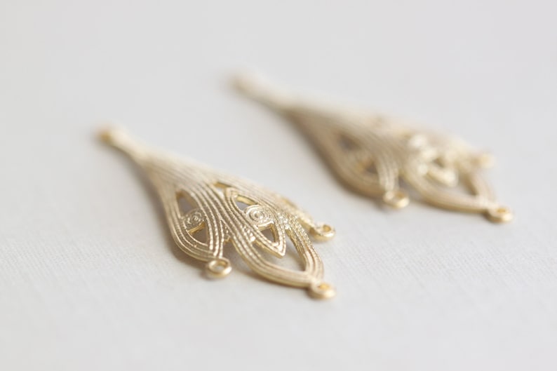 May include: Two gold-colored metal filigree embellishments with a teardrop shape and intricate designs. The embellishments have a loop at the top for attaching to a project.