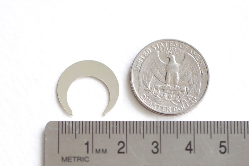 May include: A silver metal crescent moon charm, approximately 1 cm in diameter, next to a US quarter for size comparison.