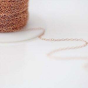 Op de afbeelding: Een close-up van een spoel met ros&eacute;gouden ketting. De ketting is delicaat en heeft een fijn, gekoppeld patroon. De ketting ligt op een wit oppervlak.