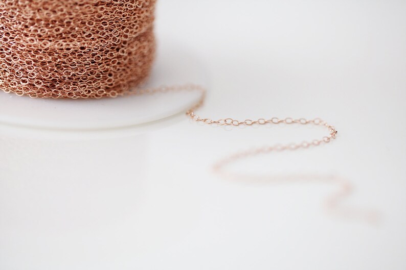 Op de afbeelding: Een close-up van een spoel met ros&eacute;gouden ketting. De ketting is delicaat en heeft een fijn, gekoppeld patroon. De ketting ligt op een wit oppervlak.