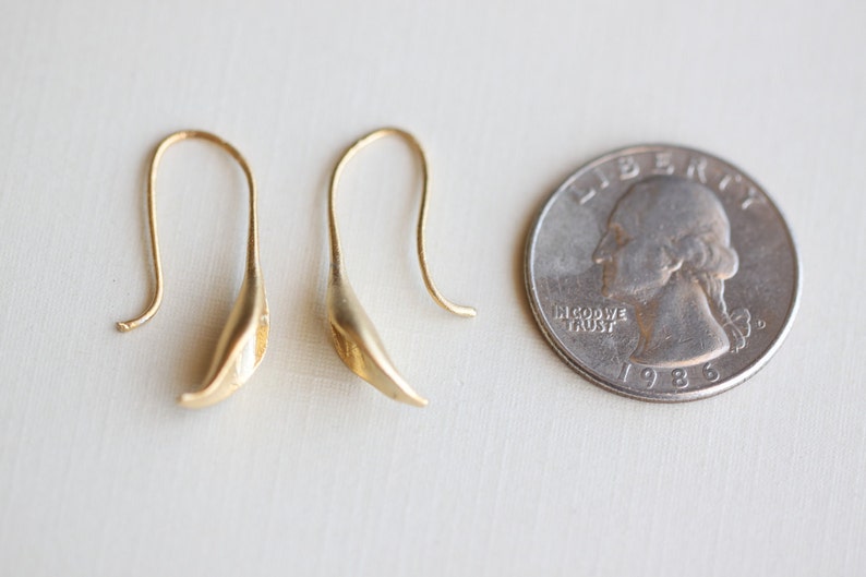 May include: A pair of gold-colored, leaf-shaped earrings with curved ear wires. A U.S. quarter coin is next to the earrings for size comparison. The coin is dated 1986.