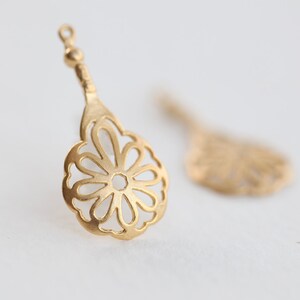 May include: A pair of gold-tone metal earring charms with a delicate, openwork flower design. The charms are shaped like a teardrop and have a loop at the top for attaching to earrings.