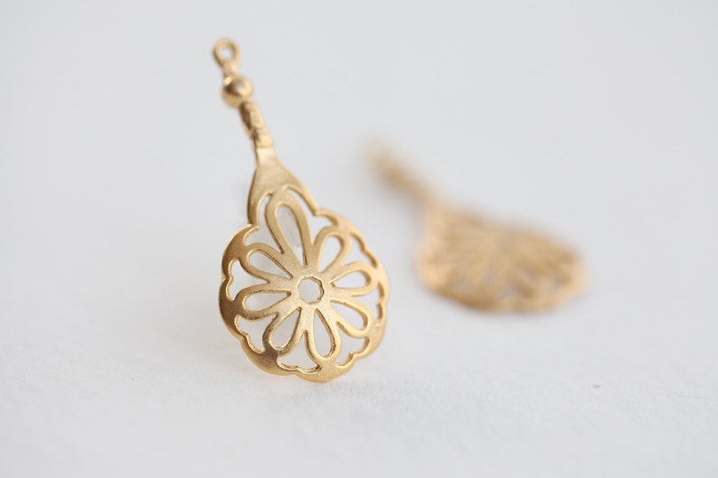 May include: A pair of gold-tone metal earring charms with a delicate, openwork flower design. The charms are shaped like a teardrop and have a loop at the top for attaching to earrings.