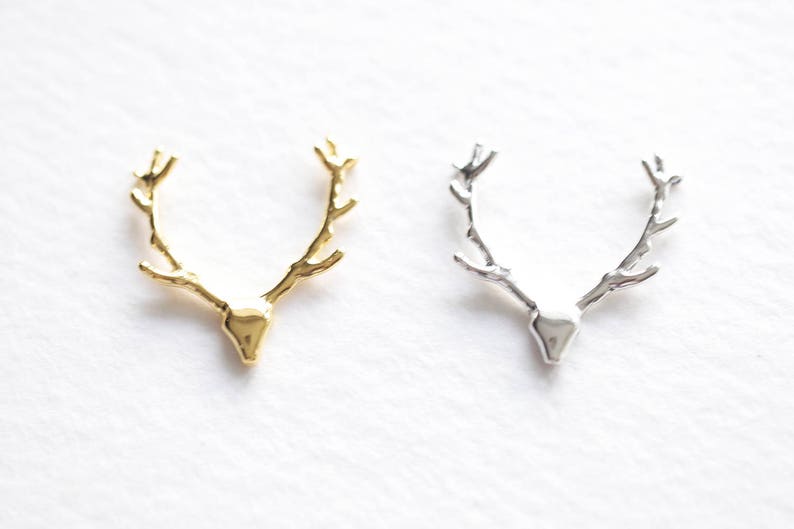 May include: Two metal deer antler pendants, one gold-toned and one silver-toned, are displayed against a white background. The pendants are detailed with a textured finish, showcasing the shape of a deer's antlers.