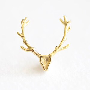May include: A gold-colored, antler-shaped ring. The ring features a detailed design of deer antlers, with a small, triangular head shape at the base. The ring is set against a white background.