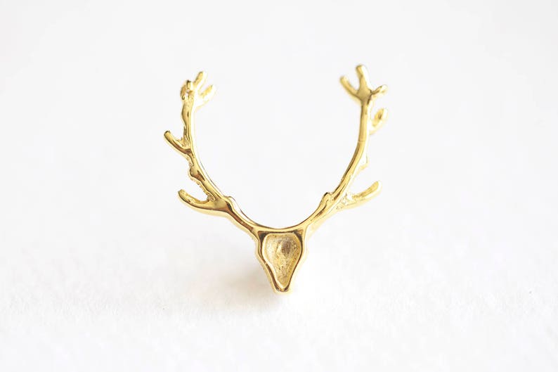 May include: A gold-colored, antler-shaped ring. The ring features a detailed design of deer antlers, with a small, triangular head shape at the base. The ring is set against a white background.