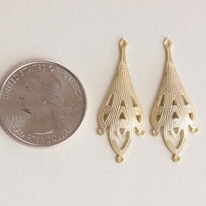 May include: Two gold-toned metal earring charms with a delicate, intricate design. The charms are shaped like teardrops with a scalloped edge and a series of concentric circles. The charms are about the size of a US quarter.